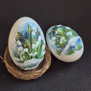 Op de afbeelding: Twee versierde paaseieren met een bloemmotief. De eieren zijn beschilderd met een blauwe achtergrond en witte bloemen met groene stelen. Eén ei rust in een klein geweven nest. De eieren zijn versierd met glitter.