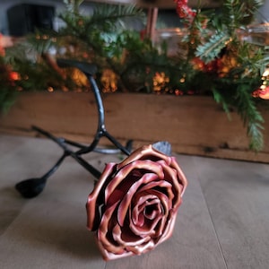 Può includere: Una scultura di rosa in metallo realizzata a mano con finitura ramata. La rosa è in piena fioritura, con petali dettagliati. Un supporto in metallo nero sostiene la rosa. Lo sfondo include vegetazione e una fioriera in legno.