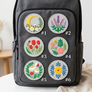 May include: A dark gray backpack featuring six circular patches with floral and celestial designs. The patches are numbered #1 through #6 and display motifs including a yellow crescent moon, lavender, red tulips, assorted colorful flowers, and daisies.