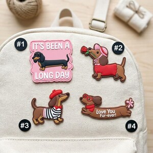 May include: Four decorative rubber patches featuring dachshunds attached to a cream-colored canvas backpack. The patches include a pink square with the text 'IT'S BEEN A LONG DAY', a dog with a rose, a dog in a beret, and a dog with the text 'Love You Fur-ever!'