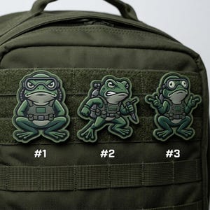 May include: Three green frog patches with military-style gear are displayed on a green backpack. Each patch features a different frog design, with the numbers #1, #2, and #3 below each frog.