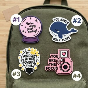 May include: Four decorative pins attached to an olive green backpack. Pin 1 is a pink crystal ball with the text "You're doing great darling!". Pin 2 is a dark blue whale with the text "YOU NEVER WALK ALONE". Pin 3 is a lightbulb with the text "MY MINDSET IS MY MOST POWERFUL TOOL". Pin 4 is a pink camera with the text "YOU ARE THE FOCUS".