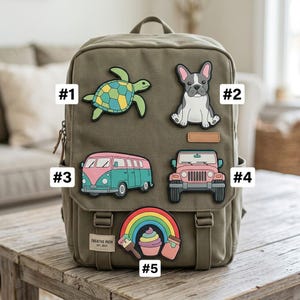 May include: An olive green canvas backpack featuring five rubberized patches: a sea turtle, a French bulldog, a pink and teal van, a pink off-road vehicle, and a rainbow with a paintbrush, cupcake, and jar. A label reads Creative Path Est. 2023.