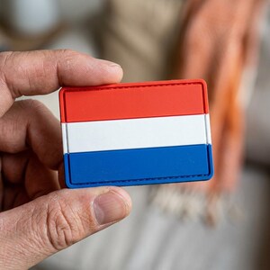 May include: A rectangular patch featuring the flag of the Netherlands. The patch has three horizontal stripes: red on top, white in the middle, and blue at the bottom. The patch is held in a person's hand.