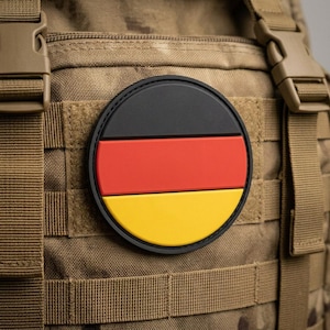 May include: A round patch featuring the German flag, with black, red, and yellow horizontal stripes. The patch is affixed to a tan tactical vest with multiple straps and buckles.