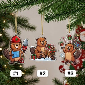 May include: Three clear acrylic Christmas ornaments featuring cartoon beavers. Ornament #1 shows a beaver with a red hard hat, blue blueprints, and a shovel. Ornament #2 shows a beaver with a wheelbarrow of gifts. Ornament #3 shows a joyful beaver.