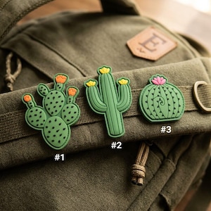 May include: Three green cactus-shaped shoe charms labeled #1, #2, and #3. Charm #1 is a prickly pear with orange flowers, #2 is a tall saguaro with yellow flowers, and #3 is a round barrel cactus with a pink flower, all featuring black details.