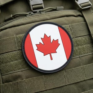 May include: A round patch featuring the Canadian flag, with a red maple leaf on a white background, flanked by red vertical stripes. The patch is bordered by a black ring and is affixed to an olive green tactical backpack.