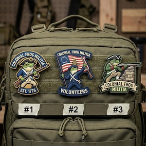 May include: Three embroidered patches featuring a frog in colonial attire. The patches are labeled #1, #2, and #3. The patches have the text "COLONIAL FROG MILITIA" and "EST. 1776", "VOLUNTEERS", and "READY AYE READY". The patches are on a green backpack.