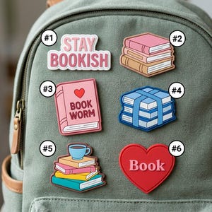 May include: Six decorative pins attached to a sage green backpack. The pins feature a pink 'STAY BOOKISH' sign, a stack of three books, a pink book labeled 'BOOK WORM' with a heart, a stack of blue books, a stack of books with a cup, and a red heart.