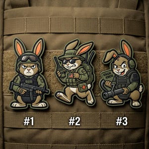 May include: Three cartoon bunny patches in military attire. Each bunny is a different design, wearing a helmet, sunglasses, and headphones. The patches are numbered #1, #2, and #3. The background is a tan tactical vest.