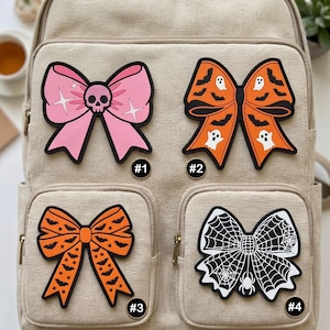 May include: Four decorative bow patches attached to a beige canvas backpack. Patch #1 is pink with a skull center. Patch #2 is orange with black bats and white ghosts. Patch #3 is orange with black bats. Patch #4 is black and white with a spiderweb pattern.