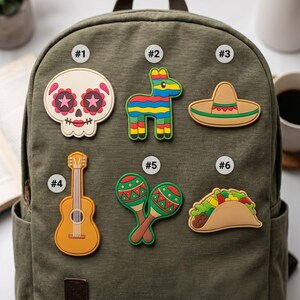 May include: Six colorful rubber patches attached to an olive green canvas backpack. The designs include a sugar skull labeled #1, a striped piñata labeled #2, a sombrero labeled #3, a guitar labeled #4, two maracas labeled #5, and a taco labeled #6.