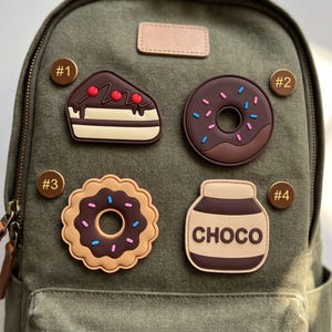 May include: Four food-themed silicone charms attached to an olive green canvas backpack. The charms include a slice of chocolate cake, a round chocolate donut with sprinkles, a scalloped chocolate cookie, and a jar labeled CHOCO.