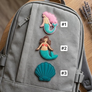 May include: A gray backpack featuring three decorative patches: #1 is a mermaid with pink hair and a teal tail, #2 is a mermaid with brown hair and a teal tail, and #3 is a teal seashell. Each patch is clearly labeled with its corresponding number.