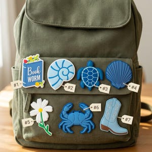 May include: An olive green canvas backpack decorated with seven rubber charms labeled #1 through #7. The charms include a blue book labeled Book Worm, a light blue nautilus shell, a blue sea turtle, a dark blue seashell, a white daisy, a blue crab, and a light blue cowboy boot.