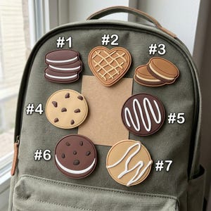 May include: Seven cookie-shaped silicone charms attached to an olive green backpack. The charms include a sandwich cookie, a heart-shaped waffle, a round cookie with chocolate chips, a dark cookie with white drizzle, and other assorted cookie designs.