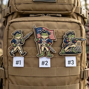 May include: Three embroidered patches featuring a frog dressed as a Revolutionary War soldier. The patches are numbered #1, #2, and #3. The frog in patch #2 is holding a flag. The patches are attached to a tan backpack.