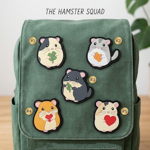 May include: A green canvas backpack featuring five hamster-themed patches labeled #1 through #5. The text THE HAMSTER SQUAD is printed above. The patches depict hamsters holding items like a four-leaf clover, orange snacks, a green leaf, and red hearts.