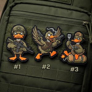 May include: Three embroidered patches featuring cartoon ducks in military-themed poses. One duck wears a helmet and holds a rifle, another holds two pistols, and the third is on a bomb with a grenade and spatula. Each patch has a number.