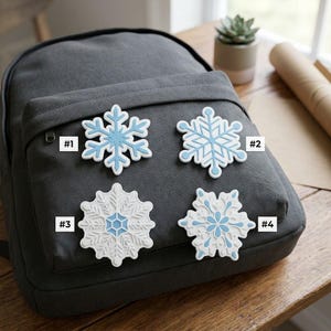 May include: Four distinct snowflake-shaped pins are arranged on a dark gray backpack. Each pin features a unique geometric pattern in shades of white and light blue, labeled with the numbers #1, #2, #3, and #4 next to each respective design.