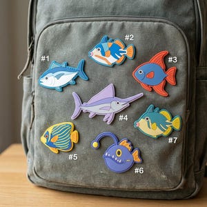 May include: Seven colorful fish-shaped silicone charms labeled #1 through #7, featuring a blue tuna, a patterned triggerfish, a red angelfish, a purple swordfish, a striped angelfish, a purple anglerfish, and a yellow-blue fish on a gray backpack.