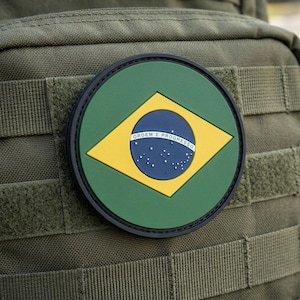 May include: A circular patch featuring the Brazilian flag, with a green background and a yellow diamond containing a blue globe. The globe has white stars and the words "Ordem e Progresso". The patch is attached to a green fabric.