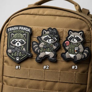 May include: Three patches featuring cartoon raccoons. One patch has the text "Trash Panda Recon" and a raccoon holding a rifle. The other two patches show raccoons with military gear, one holding a rifle and the other with a donut and coffee.