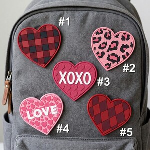 May include: Five heart-shaped patches attached to a gray backpack. Patch #1 has a red and black buffalo plaid pattern. Patch #2 features a pink and black leopard print. Patch #3 is pink with embossed hearts and the white text XOXO. Patch #4 is pink with embossed hearts and the white text LOVE. Patch #5 has a red and dark red argyle pattern.