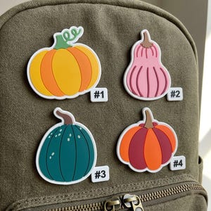 May include: Four decorative pumpkin-shaped charms attached to a green canvas backpack. The charms include an orange pumpkin labeled #1, a pink gourd labeled #2, a dark teal pumpkin labeled #3, and an orange and maroon pumpkin labeled #4.