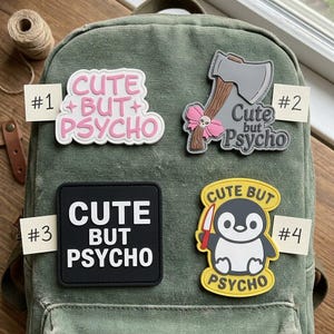 May include: Four rectangular and shaped patches attached to a green canvas backpack. Patch #1 is white with pink text reading CUTE BUT PSYCHO. Patch #2 is a grey axe with a pink bow and text. Patch #3 is black with white text. Patch #4 is a yellow penguin holding a knife.