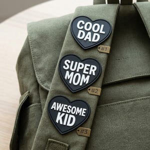 May include: An olive green canvas backpack featuring three black heart-shaped patches with white text. The patches read COOL DAD, SUPER MOM, and AWESOME KID. Each patch is accompanied by a small metallic tag labeled #1, #2, and #3 respectively.