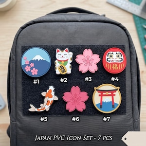 May include: A set of seven Japanese-themed PVC patches attached to a dark gray backpack. The patches feature a mountain with cherry blossoms, a beckoning cat, two pink cherry blossoms, a red Daruma doll, a koi fish, and a red torii gate.