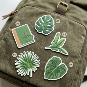 May include: Five decorative patches arranged on a green canvas backpack. The patches include a green book with a heart and coffee cup, a monstera leaf, a banana leaf, a heart-shaped leaf, and a spiky palm leaf, all with white borders.