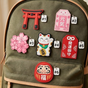 May include: An olive green canvas backpack decorated with six rubberized patches: a red torii gate, a pink omamori charm with Japanese text, a cluster of pink cherry blossoms, a beckoning cat, a red omamori charm, and a red Daruma doll with text.