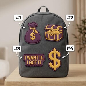 May include: A dark gray backpack featuring four decorative patches: a purple money bag with a gold dollar sign, a purple treasure chest with gold accents, a purple rectangular patch with the text I WANT IT, I GOT IT, and a large gold dollar sign.