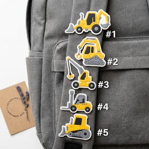 May include: Five yellow and gray construction vehicle charms labeled #1 through #5, including a backhoe, an excavator, a crane, a forklift, and a bulldozer, arranged vertically on a gray fabric backpack strap with a small card nearby.
