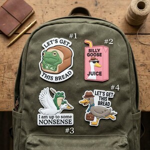 May include: Four decorative patches on an olive green backpack. Patch 1 shows a frog in bread with text "LET'S GET THIS BREAD". Patch 2 is a pink juice box labeled "SILLY GOOSE JUICE". Patch 3 shows a frog riding a goose with "I am up to some NONSENSE". Patch 4 is a goose with a briefcase and hat with "LET'S GET THIS BREAD".
