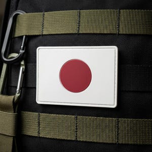 May include: A rectangular patch featuring the flag of Japan. The patch has a white background with a centered, solid red circle. The patch is attached to a black backpack with olive green straps and a metal carabiner.