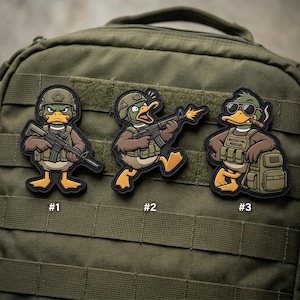 May include: Three cartoon duck patches with military themes. Each duck is wearing a helmet and tactical gear, holding a rifle. One duck is smoking a cigar and has the text "WATERFOWL WARFARE". The patches are affixed to a green backpack.