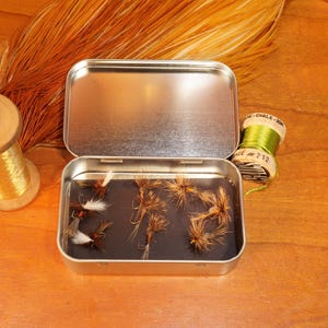 May include: A rectangular metal tin containing several brown and white feathered fishing flies, positioned next to a wooden spool of gold wire and a small spool of green thread, with a large brown feather plume fanned out in the background.