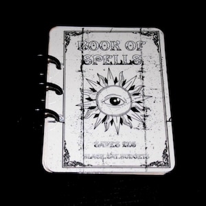 May include: A white ring-bound notebook with a black graphic print on the cover. The text reads BOOK OF SPELLS, SAVES £105, and BLACK CAT BUDGETS. A central illustration features an eye surrounded by sun rays within a decorative rectangular border.