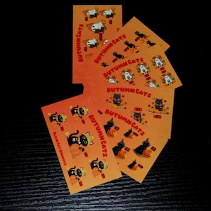 May include: A collection of rectangular orange cards labelled Autumn Cats, featuring illustrations of black cats and ghosts with pumpkins. Each card displays various price points in British pounds, such as 50p, £1, and £2, alongside small cat-themed graphics.