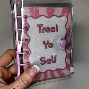 May include: A small, clear binder with a pink spine and a purple snap closure. The binder features a decorative card with the words "Treat Yo Self" in a pink and white design.