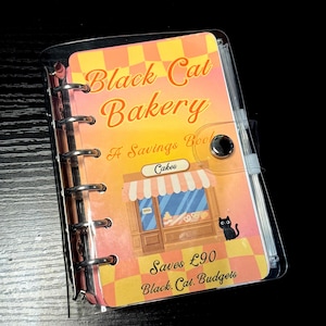 May include: A clear plastic six-ring binder featuring a cover with a yellow and orange checkered pattern. The cover displays the text Black Cat Bakery, A Savings Book, Cakes, Saves £90, and Black Cat Budgets, with an illustration of a bakery.