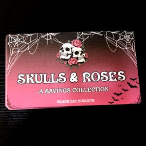 May include: A rectangular savings tracker card with a pink to black gradient background. It features white spiderwebs, two skulls surrounded by pink roses, and black bats. The text reads SKULLS & ROSES A SAVINGS COLLECTION and BLACK.CAT.BUDGETS.
