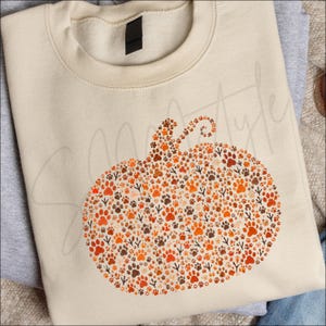 Puede incluir: Una sudadera de cuello redondo color crema con un gráfico de calabaza grande compuesto por numerosas huellas de patas y rastros de aves en tonos naranja, marrón y marrón oscuro. La sudadera presenta un cuello acanalado y una etiqueta negra.