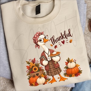 May include: A beige crewneck sweatshirt featuring a graphic of a goose wearing a red floral headscarf, glasses, and a brown cardigan. The goose holds a pie and a book with a teacher tag. Text reads Thankful, surrounded by autumn leaves and pumpkins.