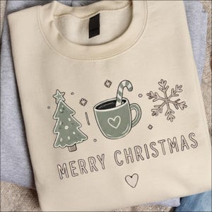 Op de afbeelding: Een beige sweater met ronde hals en een grafische print van een groene kerstboom, een groene mok met een zuurstok en een sneeuwvlok. De tekst MERRY CHRISTMAS staat onder de afbeeldingen in een eenvoudig lettertype, met een klein hartje.
