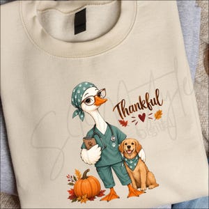 Può includere: Una felpa beige con la grafica di un'oca vestita da veterinario con camice verde acqua, occhiali e cuffia, che tiene un blocco note. Accanto all'oca c'è un golden retriever con una bandana verde acqua, una zucca e la scritta Thankful.